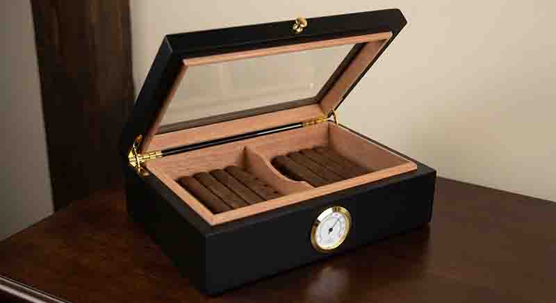 Geöffneter Humidor aus edlem Holz mit mehreren Zigarren in verschiedenen Formaten, die ordentlich gelagert sind. An der Vorderseite des Gehäuses ist ein kreisförmiges Hygrometer mit goldenem Rahmen eingelassen, dessen Nadel auf eine weiße Skala zeigt.