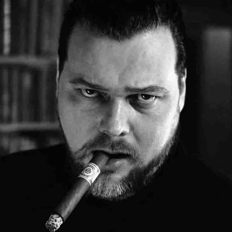 Schwarz-Weiß-Porträt zeigt einen bärtigen Mann in einer Nahaufnahme, der eine brennende Zigarre im Mund hält und mit einem intensiven, ernsten Blick direkt in die Kamera schaut. Der Hintergrund ist dunkel und unscharf, was den Fokus ganz auf sein Gesicht und den aufsteigenden Rauch lenkt.