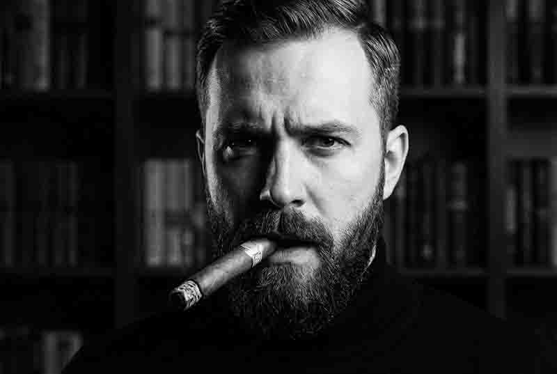 Dramatisches Schwarz-Weiß-Porträt eines Mannes mit Vollbart und Rollkragenpullover, der eine kubanische Habano raucht, während er vor einem dunklen Bücherregal direkt in die Kamera schaut.